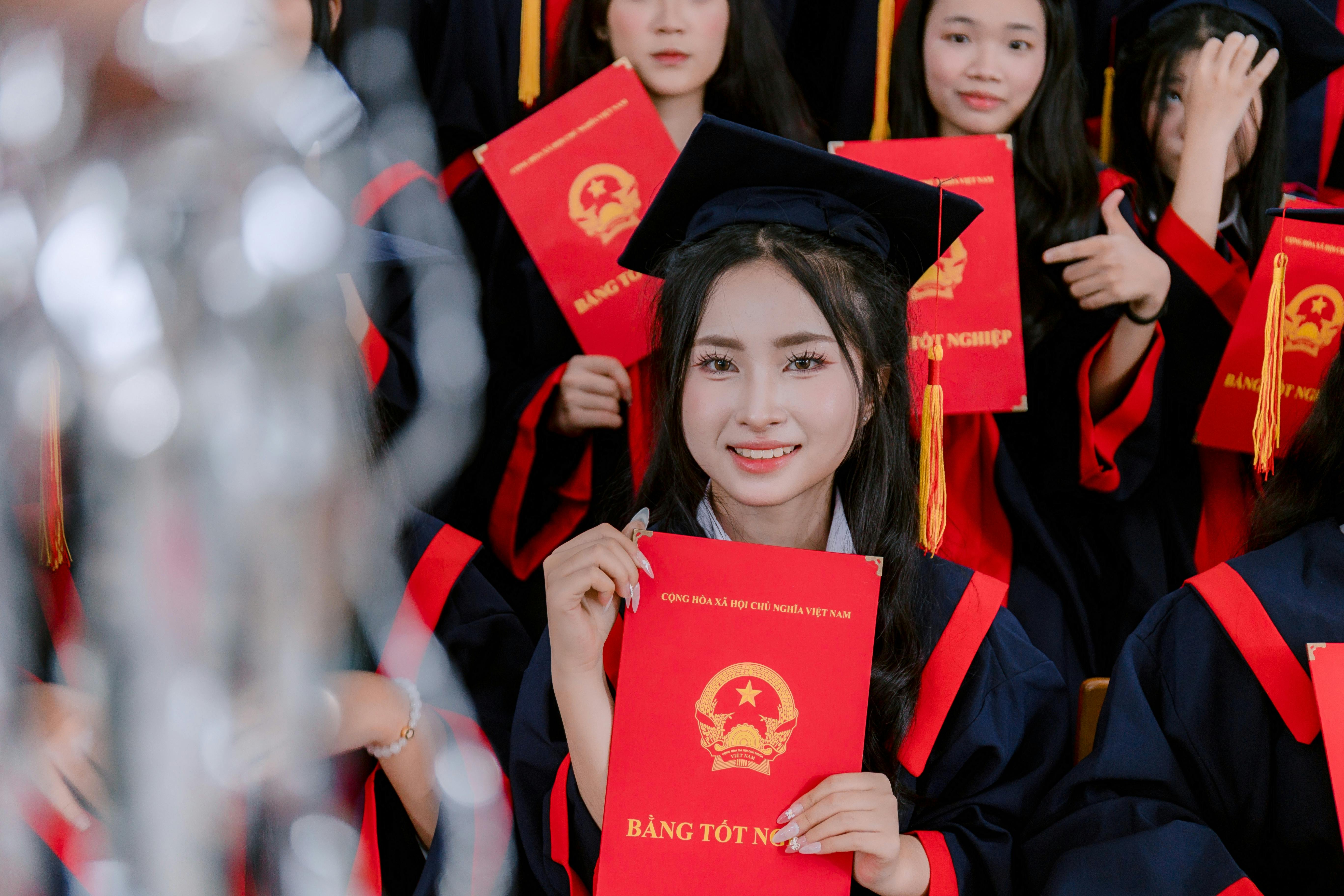 A cheerful female graduate holds a diploma during a graduation ceremony, surrounded by fellow graduates in caps and gowns.